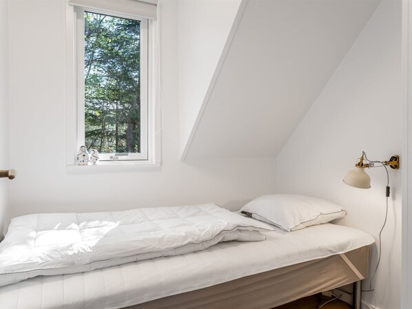 A small, white, single-bed room with a sloped ceiling, a window with a view of trees, and a wall-mounted lamp.
