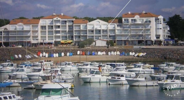 Un port de plaisance où sont amarrés de nombreux bateaux, un grand bâtiment doté de plusieurs balcons et une côte rocheuse.