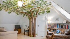 Ein gemütliches Wohnzimmer mit einer großen Baum-Wandmalerei, einem Ledersofa, einem Bücherregal und einem Fernseher.