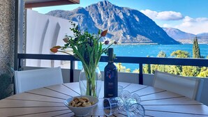 A table with a glass vase of flowers, a bottle of wine, and a bowl of snacks on a balcony with a mountain view. - Blue Vista Lugano Apt.616 by Interhome (Bissone)