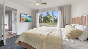 A bedroom with a bed, a television, a window with curtains, and a view of greenery outside. - The Lazy Pelican - Tea Gardens (Tea Gardens)