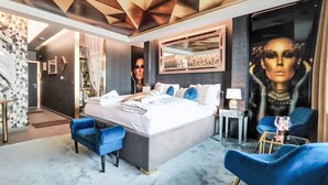 Una camera d'albergo moderna con un letto grande, due poltrone blu, un piccolo comodino e uno specchio grande.