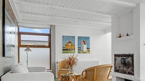 A living room with a white sofa, a wicker chair, a glass coffee table, and a fireplace. There are two paintings on the wall and a window with a view of a building. - "Talida" - 500m from the sea by Interhome (Skagen)