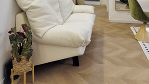 Ein modernes Wohnzimmer mit einer Couch, einem grünen Sessel und einem kleinen Tisch mit einer Pflanze. Im Hintergrund ist eine Küche zu sehen.
