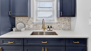 Een keuken met donkerblauwe kastjes, een wit aanrecht, een roestvrijstalen spoelbak en een betegelde achterwand, inclusief een raam met zonwering.