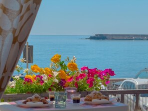 Une table avec des croissants et un verre d’eau, un vase de fleurs et une vue sur la mer.