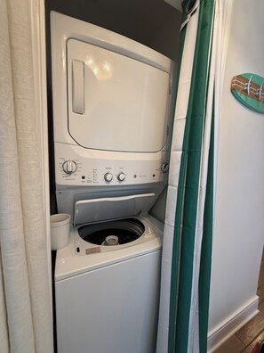 A compact washer and dryer unit with a control panel, a small storage area, and a green-striped curtain. - Brand new Boardwalk reno, king bed + bunks, perfect for couple or young family! (Panama City Beach)