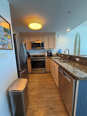A compact kitchen with wooden cabinets, a stainless steel refrigerator, a microwave, an oven, a dishwasher, and a granite countertop. - Brand new Boardwalk reno, king bed + bunks, perfect for couple or young family! (Panama City Beach)