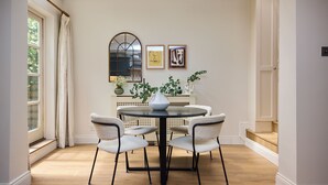 Un comedor con una mesa redonda, seis sillas y un jarrón. Hay dos cuadros en la pared, una ventana con cortinas y una escalera que lleva a un nivel superior.