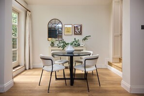 A dining area with a round table, six chairs, and a vase. There are two framed pictures on the wall, a window with curtains, and a staircase leading to an upper level. - Host My Nest: Spacious 4BR House with Garden (London)
