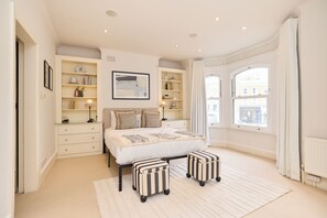 A bedroom with a large bed, two bedside tables, a bookshelf, and a window with curtains. - Host My Nest: Spacious 4BR House with Garden (London)