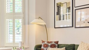 Un salón con un sofá verde, una mesa de centro redonda y una lámpara de pie. Hay cuadros enmarcados en la pared y una ventana con contraventanas.