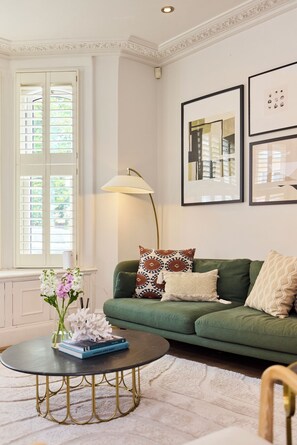 A living room with a green sofa, a round coffee table, and a floor lamp. There are framed pictures on the wall and a window with shutters. - Host My Nest: Spacious 4BR House with Garden (London)