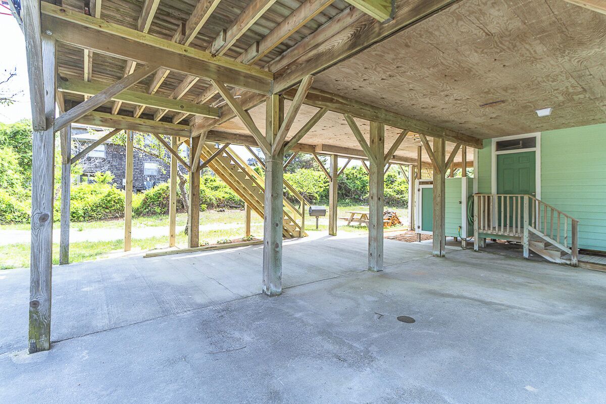 Un pavillon ouvert doté d’un plancher en béton et de poutres en bois, d’une porte verte et d’un escalier menant à une zone abritée.