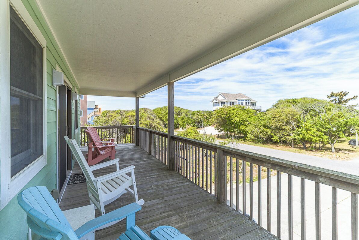 A covered porch with two blue Adirondack chairs, a wooden railing, and a view of a house and trees.