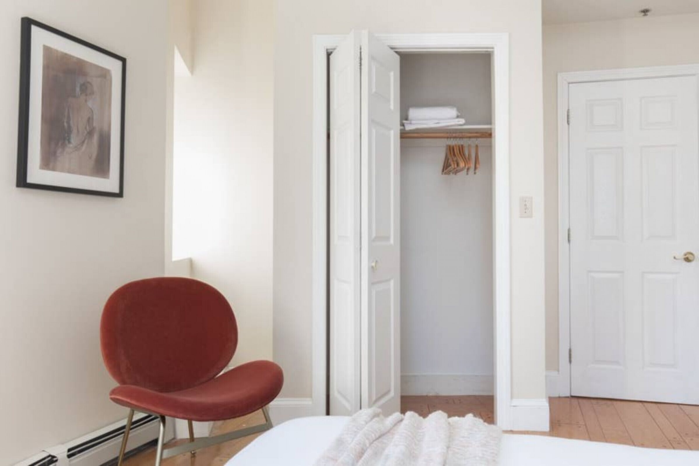 A bedroom with a bed, a red chair, a white door, and a closet with hanging clothes.