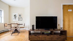 A living room with a wooden entertainment unit, a flat-screen TV, and a dining area with a round table and chairs. - Walk everywhere - Seaport, North End, Beacon Hill.  Steps from MBTA public transit.  Keyless Entry! (Boston)
