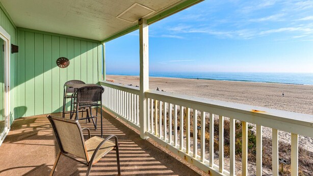 Een overdekt terras met twee rieten stoelen en een tafel, met uitzicht op een strand en een heldere lucht.