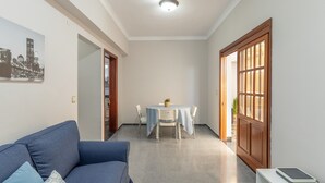 Una habitación con un sofá azul, una mesa de comedor con sillas, una pared blanca con una pintura de paisaje urbano y una puerta de madera.