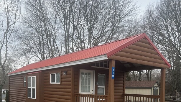 Een klein blokhut met een rood dak, een veranda en een bord met het nummer 119.