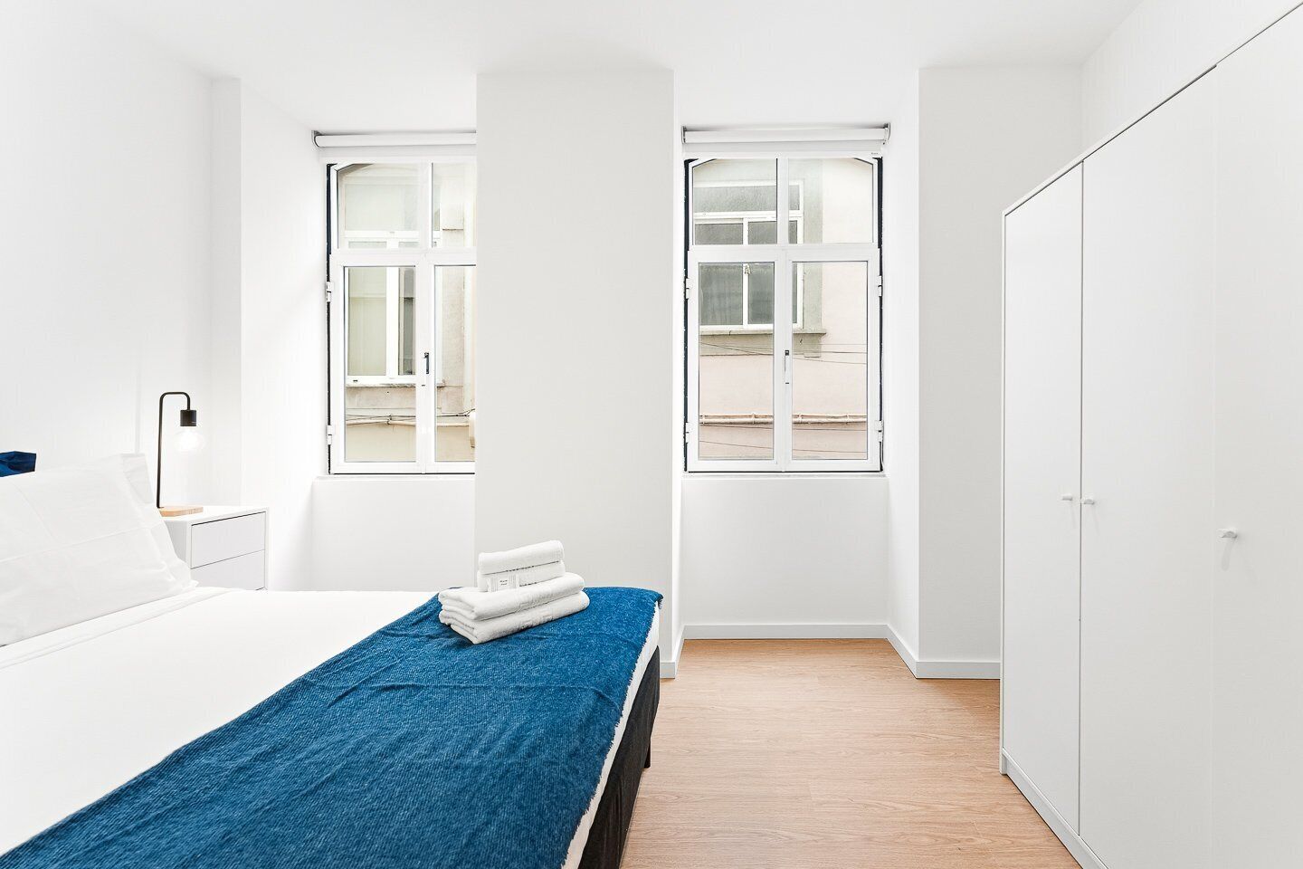 Een minimalistische slaapkamer met een bed, nachtkastje, lamp, raam en kledingkast.