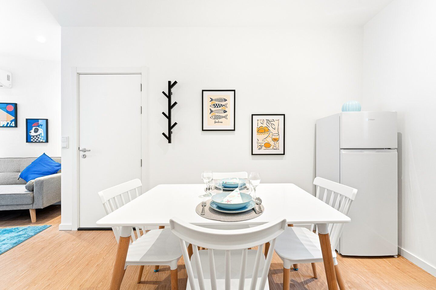 Een modern, minimalistisch eetgedeelte met een witte tafel gedekt voor vier personen, omringd door witte stoelen. Er hangen kunstwerken in lijsten aan de muur, en er is een witte deur en een witte koelkast ernaast.