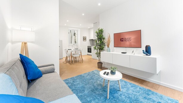 A modern living room with a grey sofa, blue cushions, a white coffee table, and a blue rug. A flat-screen TV is mounted on the wall, and a plant is on the floor. - GuestReady - The Olives - Blue (Coimbra)