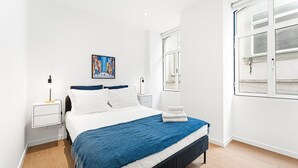 A modern hotel room with a bed, a nightstand, a bedside table, a window, and a painting on the wall. - GuestReady - The Olives - Blue (Coimbra)