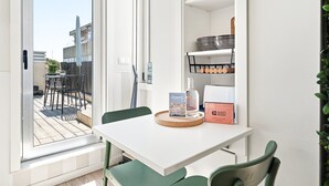 Ett rum med ett vitt bord, gröna stolar, ett fönster med utsikt över byggnader, en hylla med köksredskap och en skylt med texten ”klart för gäster”.