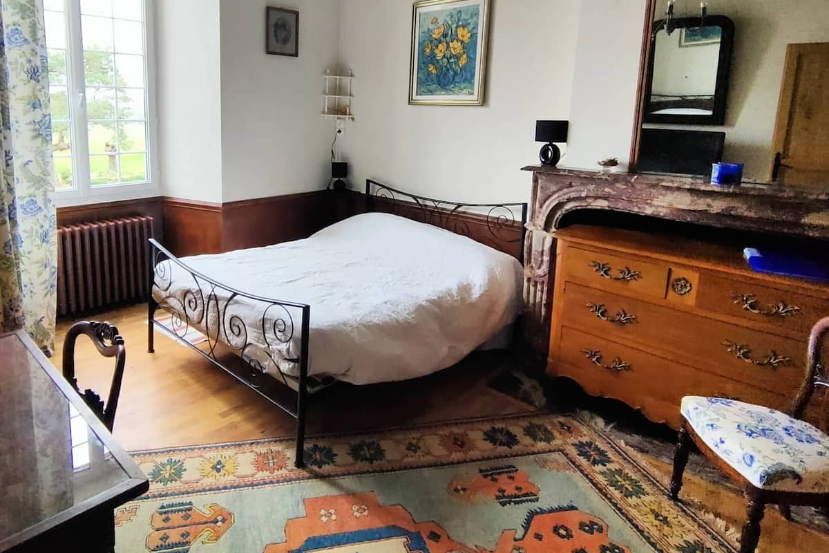 A bedroom with a bed, a dresser, a chair, a radiator, a chandelier, and a window.