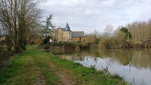 A riverside view of a historic stone building with a tower, surrounded by trees and a calm water body. - La Provosterie (Ahuillé)