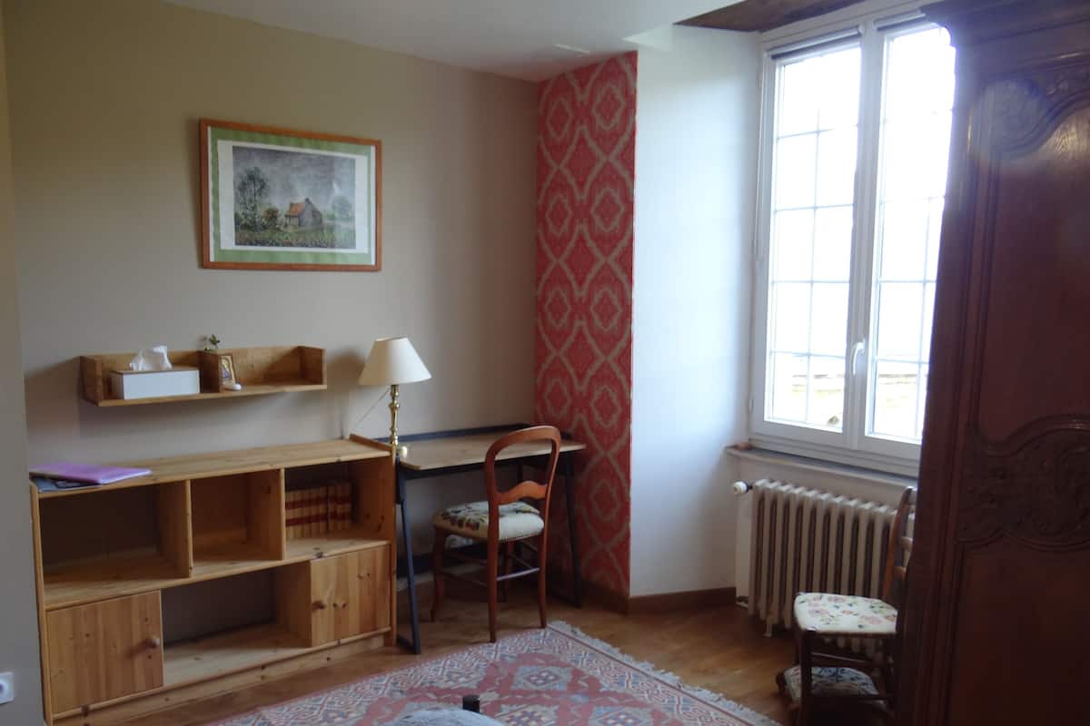 A room with a wooden desk, chair, and shelves, a radiator, a patterned rug, and a window with a view.