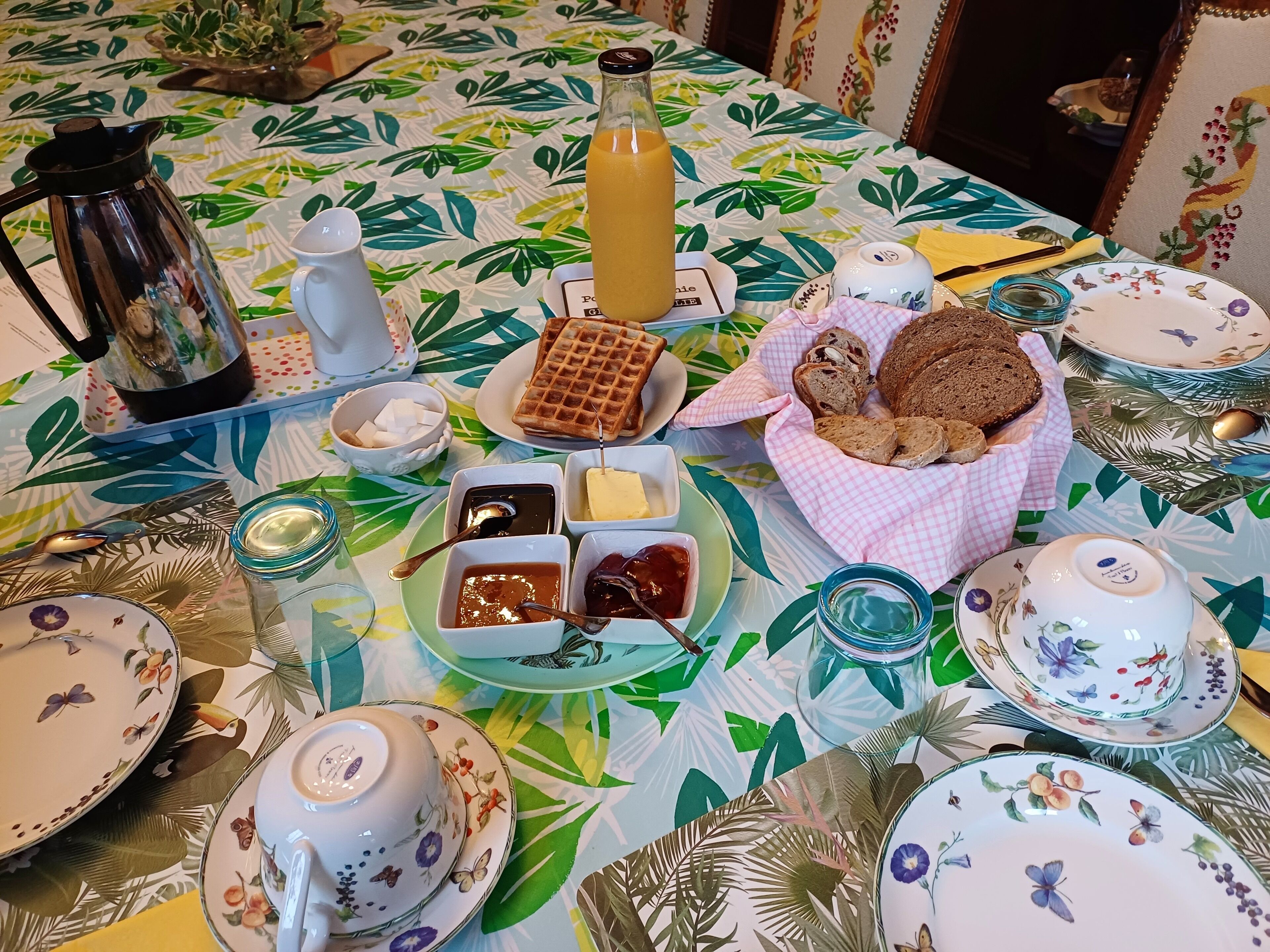 A breakfast table with a floral tablecloth, a coffee pot, a bottle of orange juice, waffles, bread, butter, jam, and tea sets.