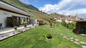 A modern building with a well-maintained lawn, outdoor seating, and a view of a vineyard and village in the background. - Zilli Studio Apartments (Termeno Sulla Strada del Vino)