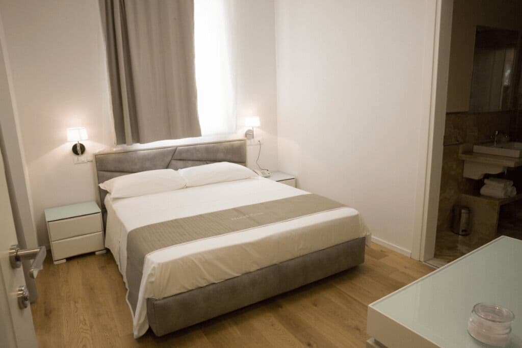 Ett hotellrum med en säng, sängbord, ett litet bord, ett glasbord och ett badrum med handfat och spegel.