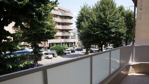 Un balcón con vista a un edificio de varios pisos, autos estacionados y árboles.