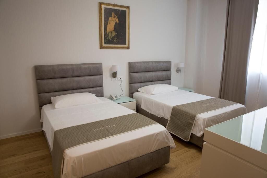 A hotel room with two beds, a bedside table with a lamp, a painting on the wall, and a window with curtains.