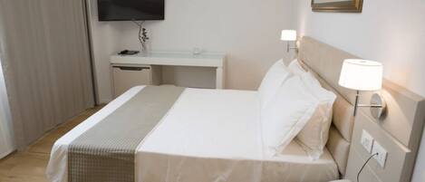 A hotel room with a bed, bedside table, lamp, phone, desk, TV, and a painting on the wall.