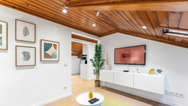 A modern hotel lobby with a wooden ceiling, a reception desk, a flat-screen TV displaying "GUEST READY", and framed artwork on the walls.