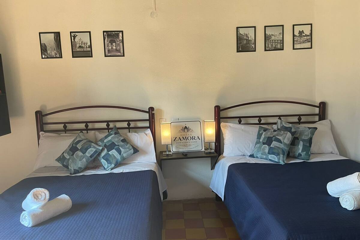 Une chambre d’hôtel avec deux lits, chacun étant équipé d’un oreiller et d’une serviette enroulée. Sur la table de chevet, on peut lire une pancarte indiquant « HÔTEL ZAMORA ».