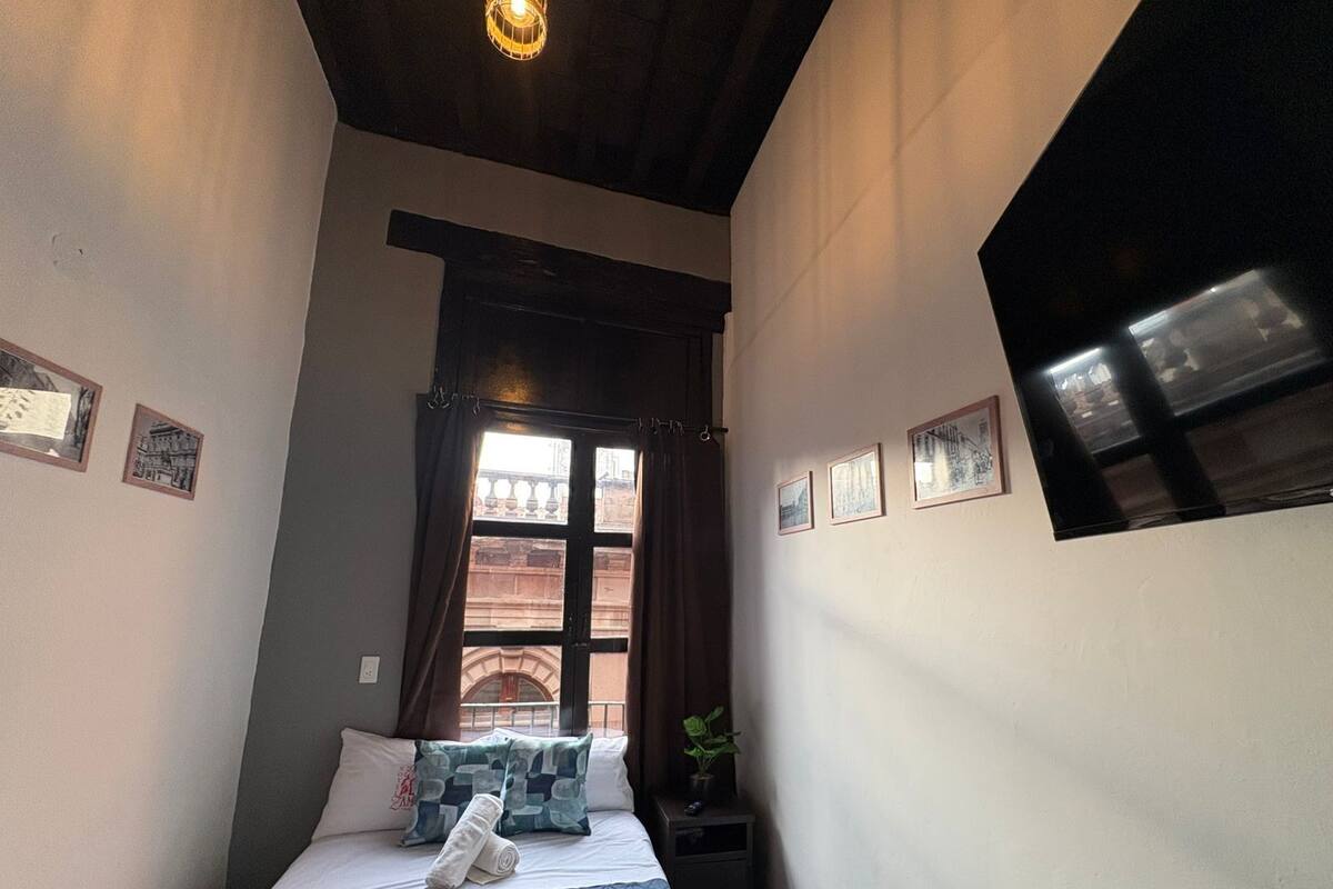 Une petite pièce agréable, avec un lit, une fenêtre avec des rideaux et un luminaire fixé au mur.