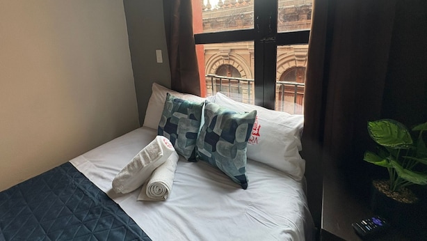 Een hotelkamer met een bed, een raam met uitzicht op een gebouw en een plant aan de rechterkant.