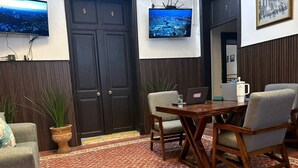 Een hal met een vloer met rood patroon, een houten tafel met stoelen, een bank, een potplant en twee wandtelevisies waarop stadsgezichten te zien zijn.