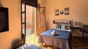 Een slaapkamer met een bed, een stoel, een nachtkastje en een raam met uitzicht op gebouwen.