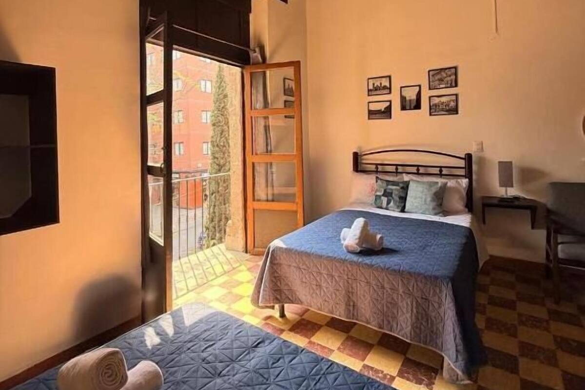 Une chambre à coucher comprenant un lit, une chaise, une table de chevet et une fenêtre donnant sur des immeubles.