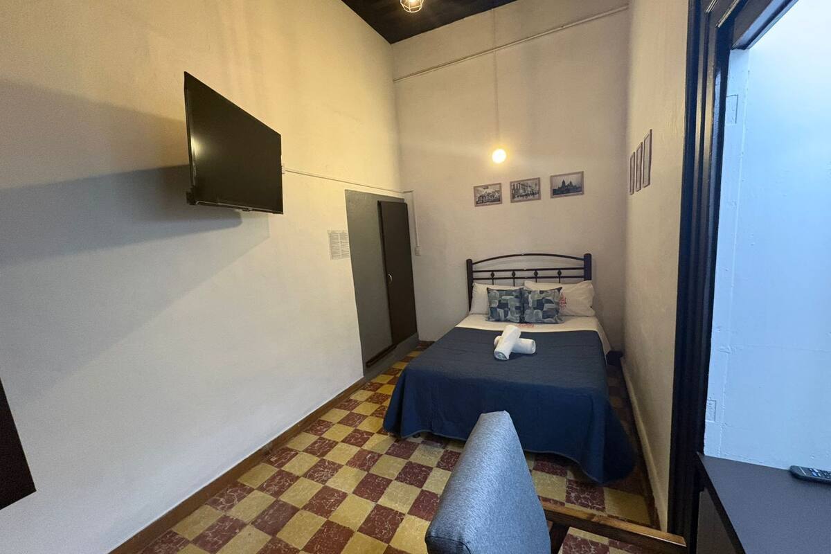 Une petite pièce comprenant un lit, une chaise, un téléviseur à écran plat fixé au mur et un sol à carreaux.