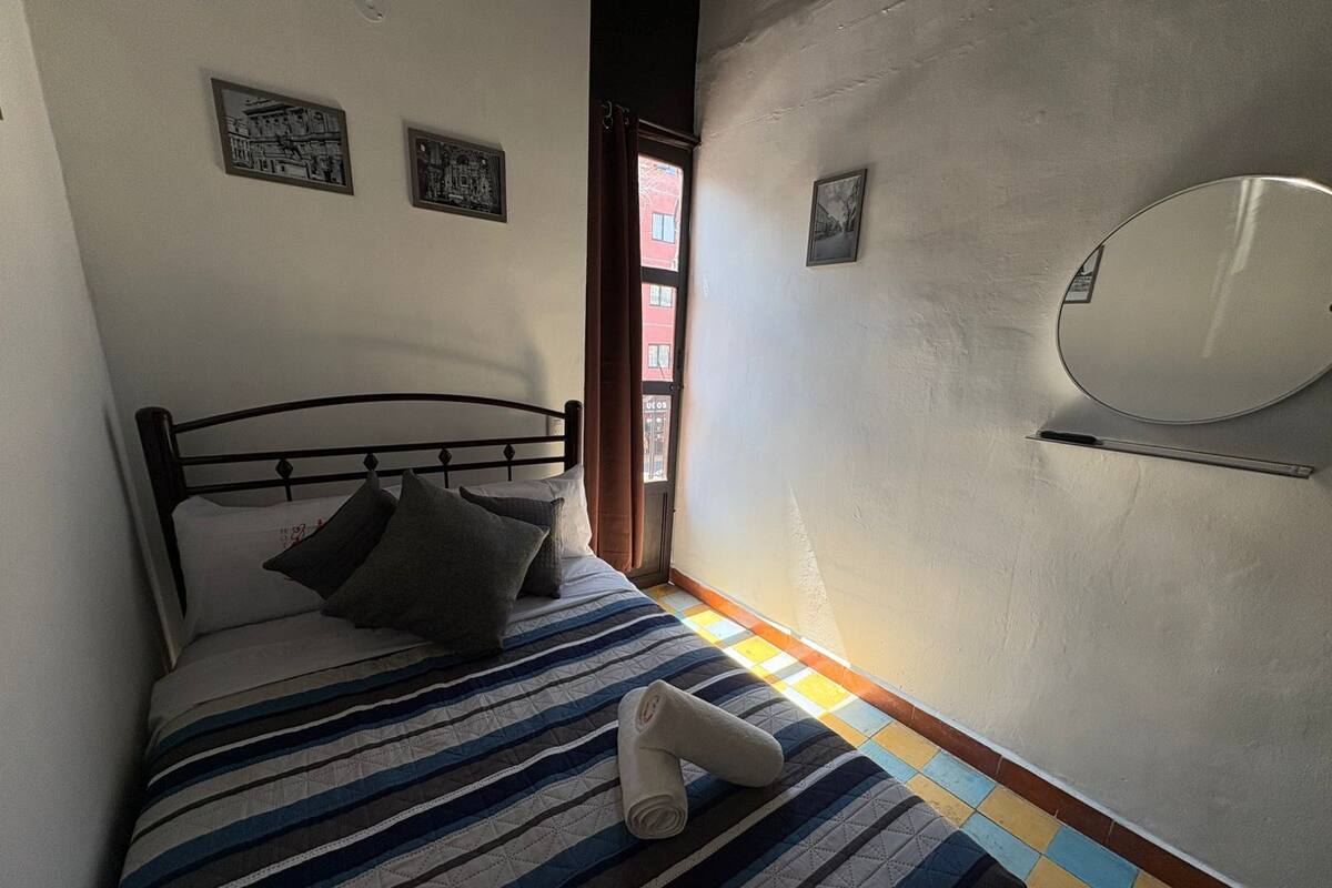 Un lit simple avec une couvre-lit à rayures, un petit miroir rond accroché au mur et une fenêtre avec des rideaux.