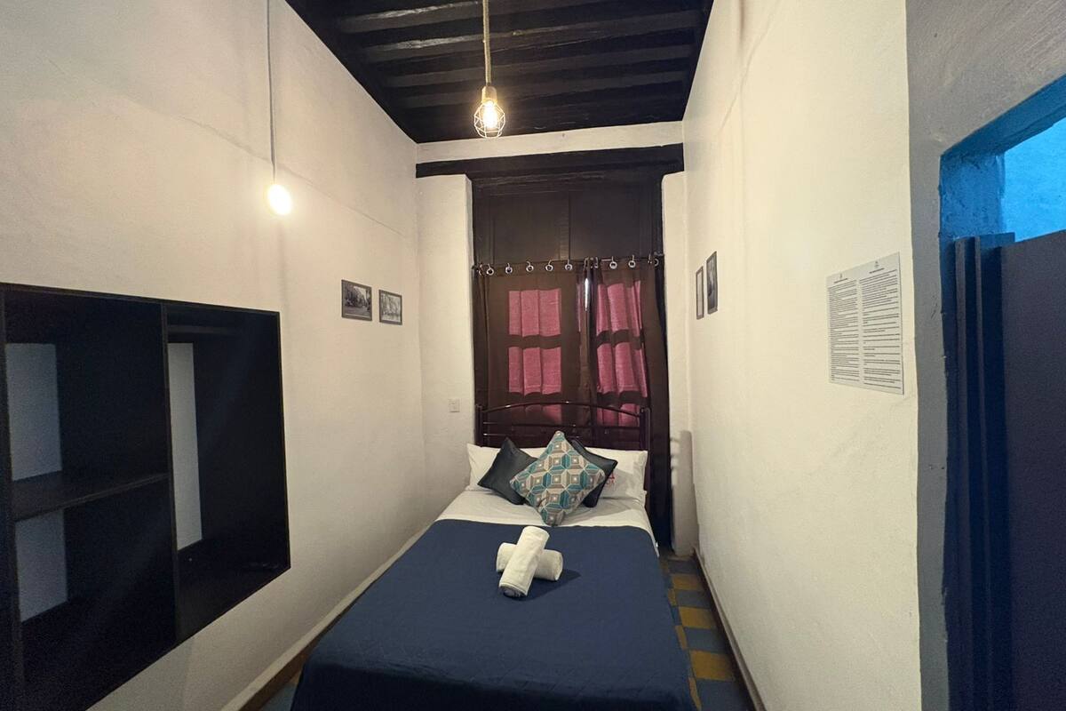 Une chambre d’hôtel étroite, avec un lit, une petite fenêtre avec des rideaux, une étagère fixée au mur et un luminaire au plafond.