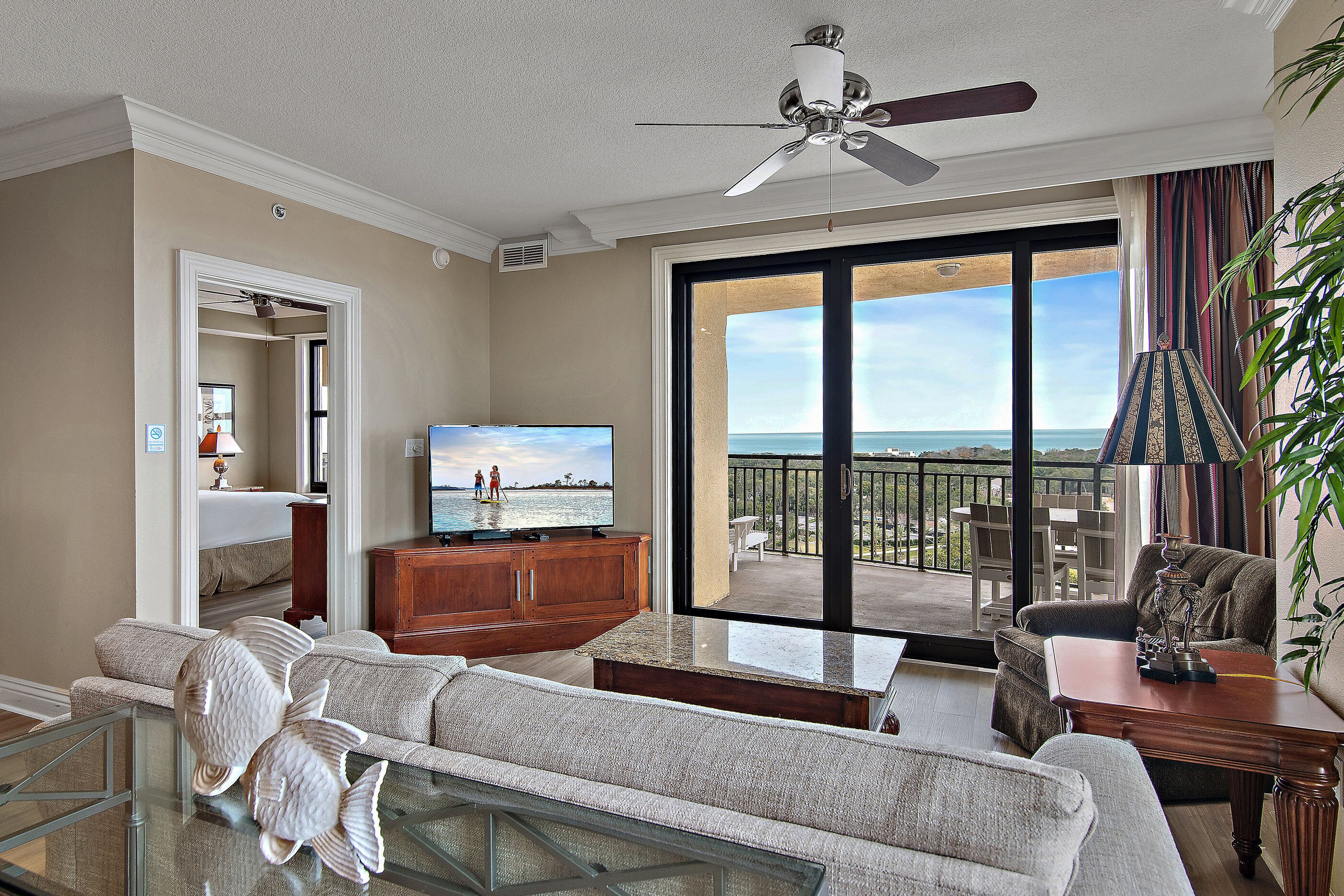 A living room with a television, a sofa, a coffee table, and a balcony with a view of the ocean.