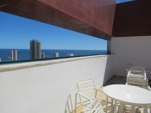 Ein Balkon mit Blick auf das Meer und Gebäude, weiße Metallstühle und ein kleiner runder Tisch.